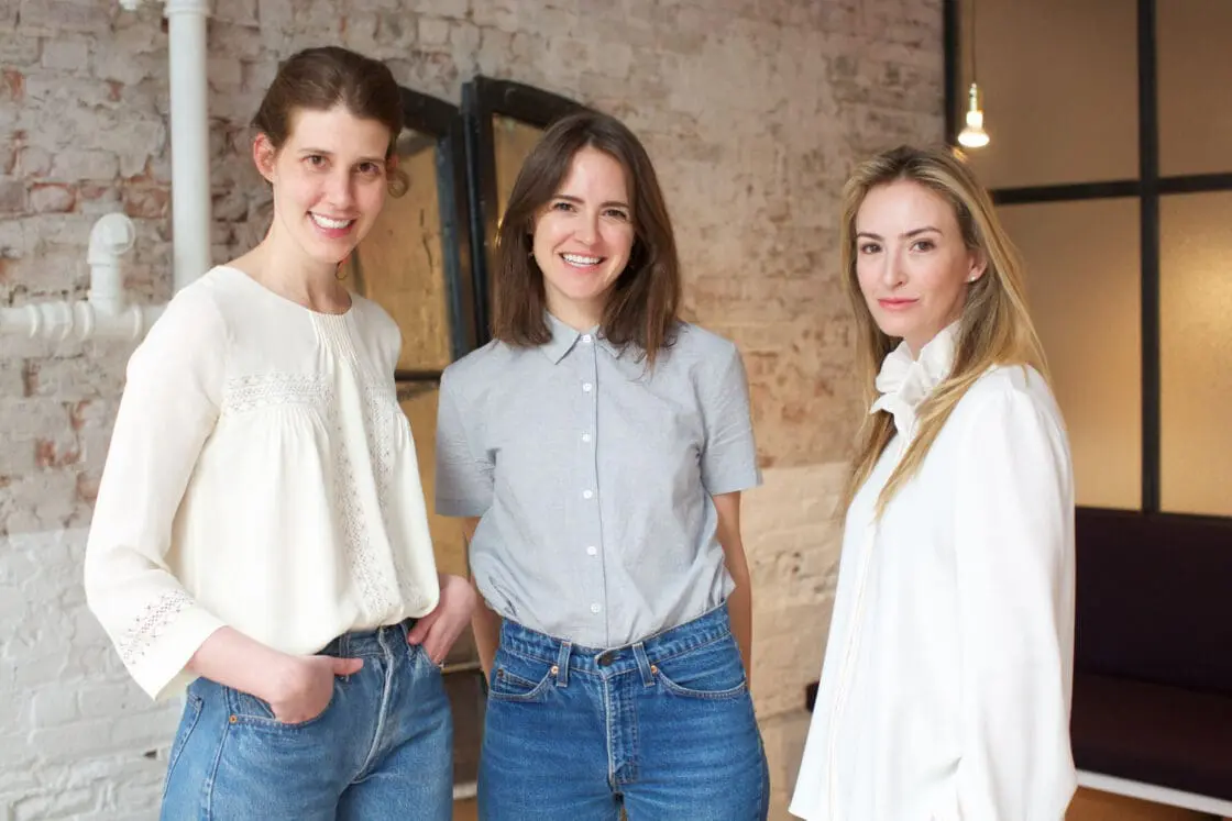 Three women in white tops and jeans standing together in a brick-walled loft