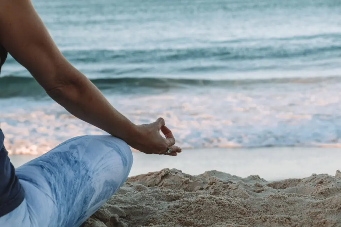 Image of a beach with a woman partially in the shot sitting cross-legged wearing yoga pants and holding hand out at her knee in gyan mudra for meditation.