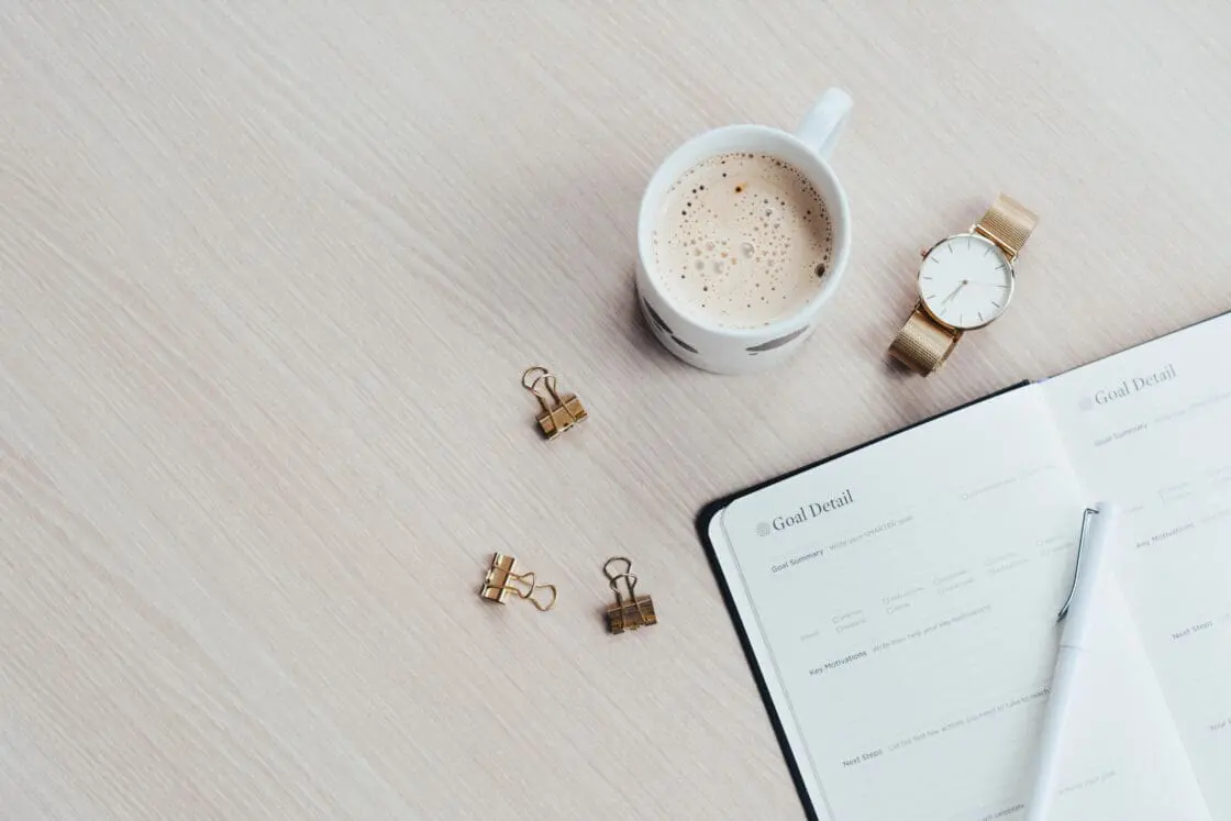 Image of a journal opened on a light wood table with a cup of coffee and a watch and three paper clips used as a tool for writing down your visualization.