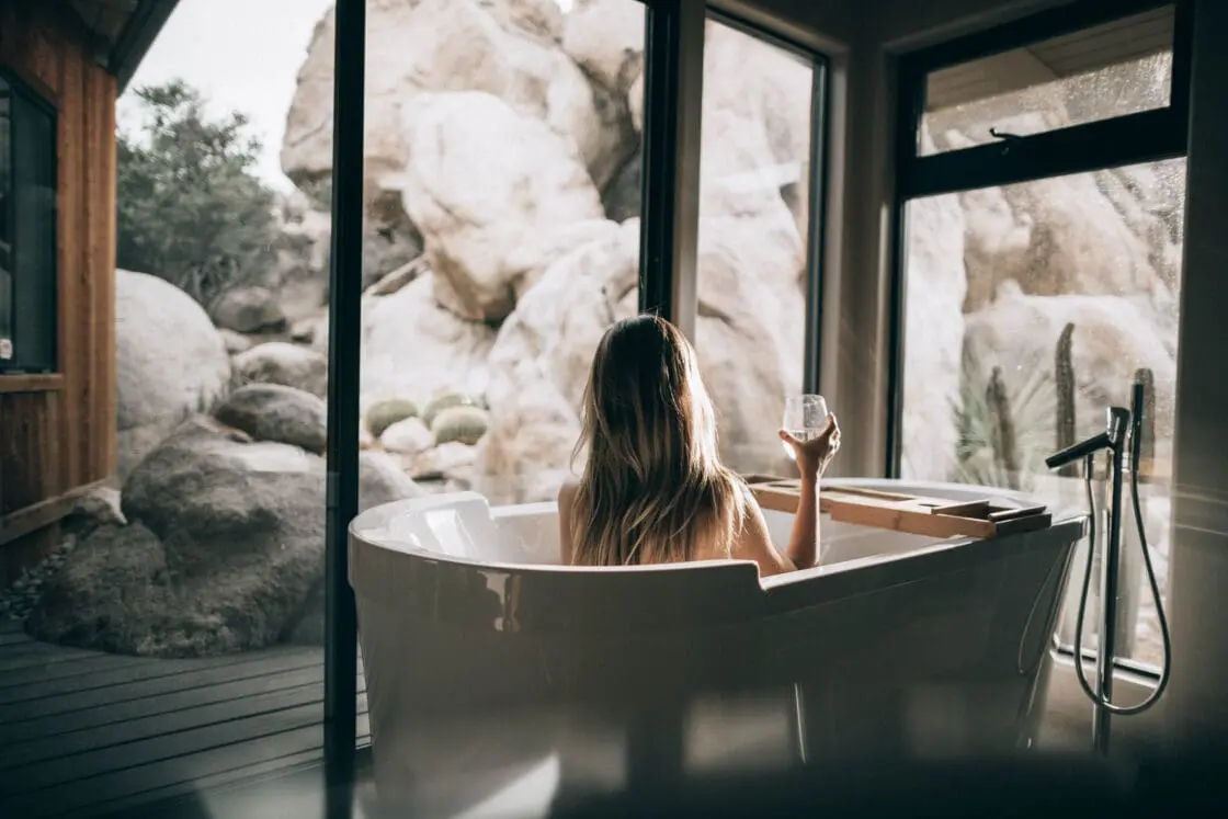 Image of the back of a woman with blond hair in a modern tub sitting upright with a glass of wine in her hands, looking out of the floor-to-ceiling windows overlooking a rocky terrain in what could be a spa, showing the importance of a calm mind when it comes to manifesting and visualization.