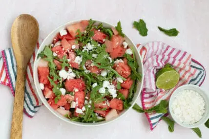 Image: watermelon feta salad with mint, arugula and feta cheese served in a white bowl on a colorful striped towel alongside a wooden spoon, halved lime, and small white bowel of feta via Organic Authrority