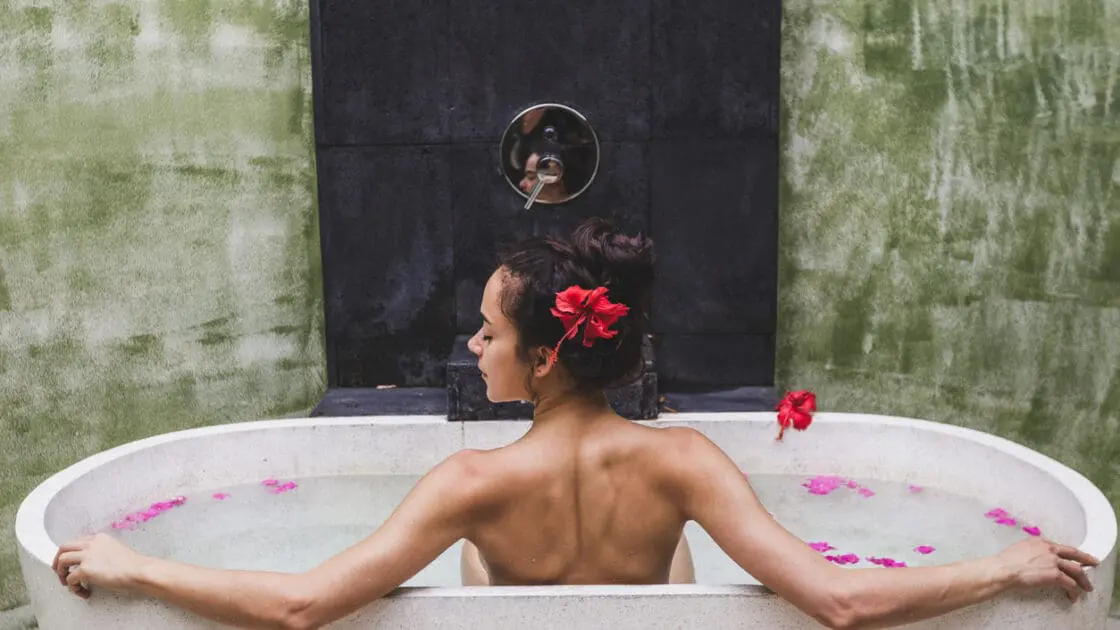 Image of a woman in a modern, stone bath, sitting up with arms splayed to each side facing a wall with bath fixture. She wears a hibiscus flower in her hair and shows the benefits of Epsom salt baths. Why to Take an Epsom Salt Bath