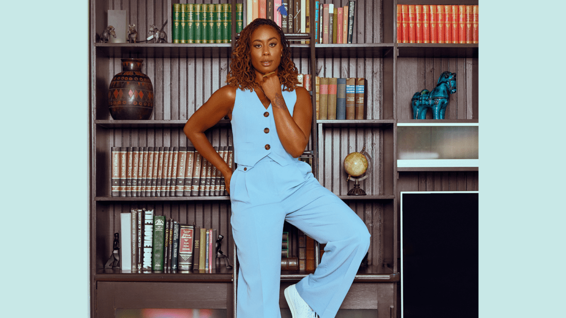 Image of actress Zuri Adele in light blue vest and slacks in front of a brown bookshelf filled with books and artifacts. She shares her passion for a personal wellness curriculum and advocates for collective liberation.