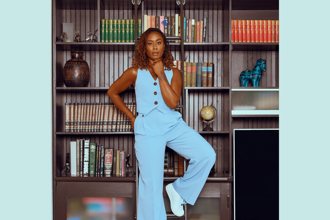 Image of actress Zuri Adele in light blue vest and slacks in front of a brown bookshelf filled with books and artifacts. She shares her passion for a personal wellness curriculum and advocates for collective liberation.