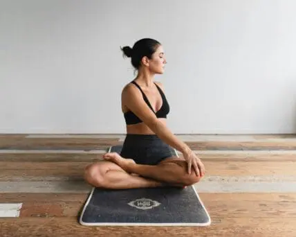 Image of a woman sitting on a black yoga mat with her legs crossed and her eyes closed as she twists to the side in a relaxing stretch.