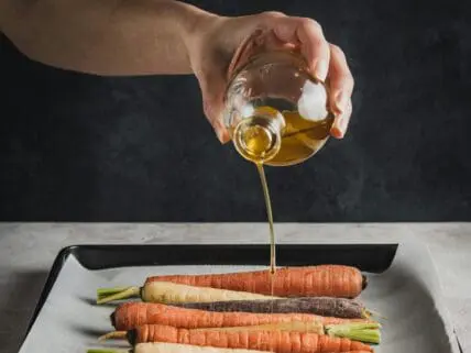 Image of a hand pouring a miscellaneous seed or vegetable oil over a tray of carrots.