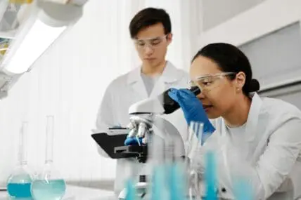 An image of a woman in laboratory wearing a lab coat, gloves, and protective goggles as she looks into a microscope. Her male coworker stands behind her taking notes on his clipboard.
