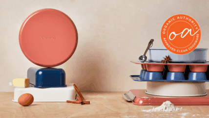 Image of the set covered in this Caraway Bakeware review, shown in mixed colors (perracotta, navy, and white) in two stacks with a single brown egg, half stick of butter, and sprinkle of flour on the wooden work surface.