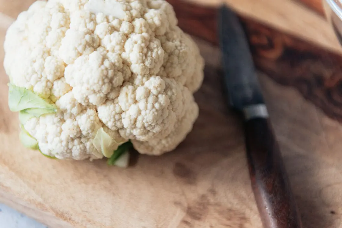 Image of a head of cauliflower sitting on a wooden chopping board beside a knife.