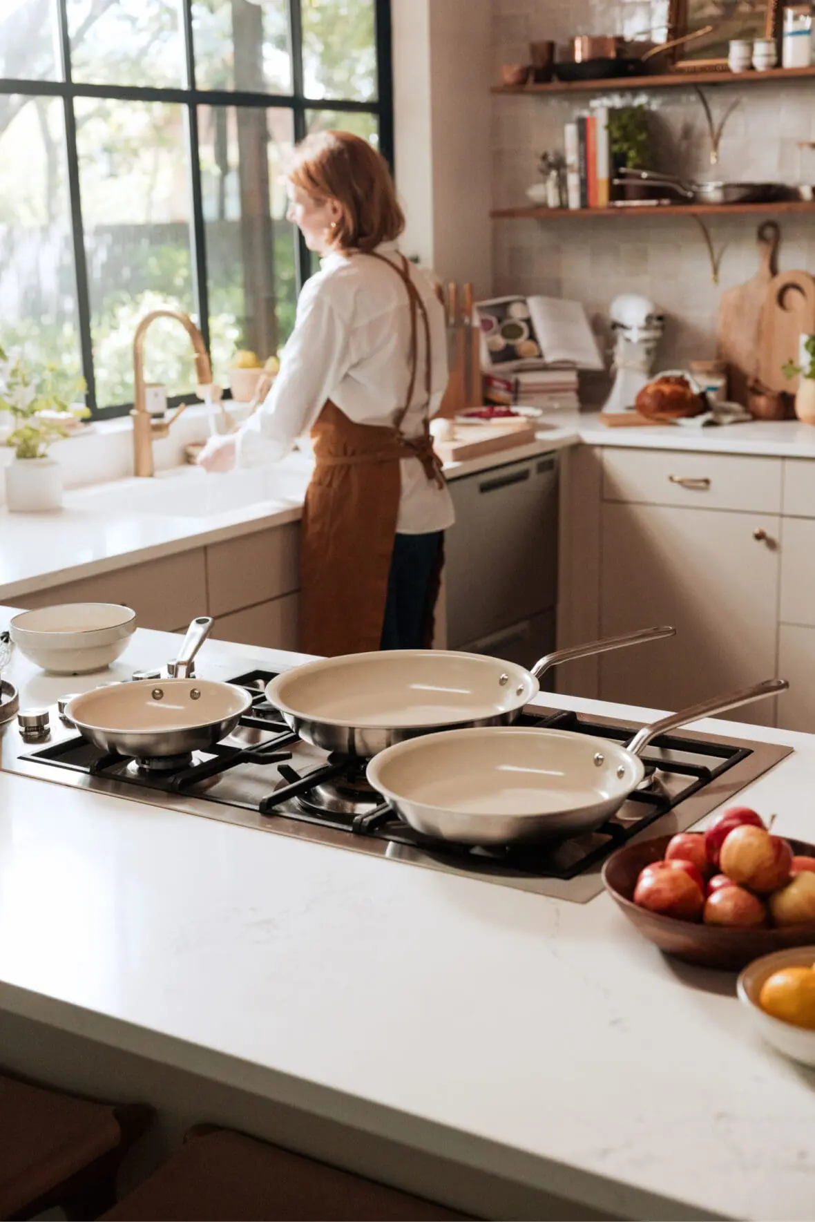 Image of a woman with an apron at a sink with a stove with clean pans in the foreground showing how easy these nonstick pans are to clean.