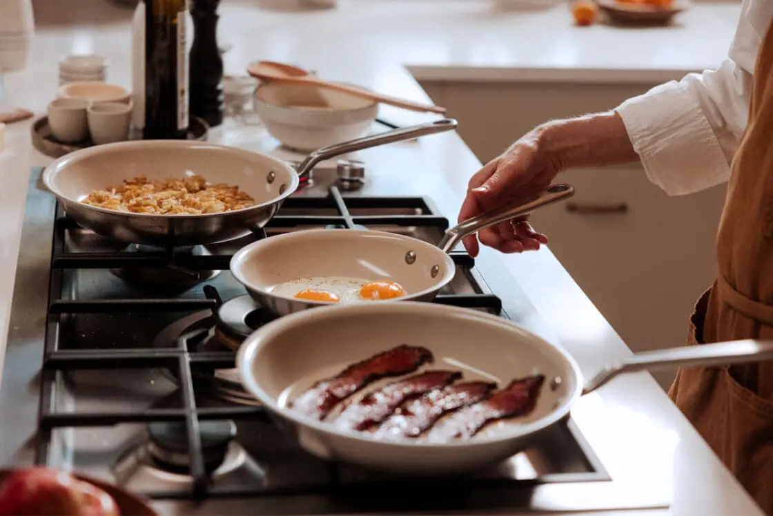 Image of three Made In CeramiClad fry pans on a stovetop, with a pair of hands maneuvering them as they cook bacon, eggs, and potatoes.