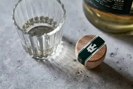 small glass of mezcal at room temp with bottle lid beside on stone surface