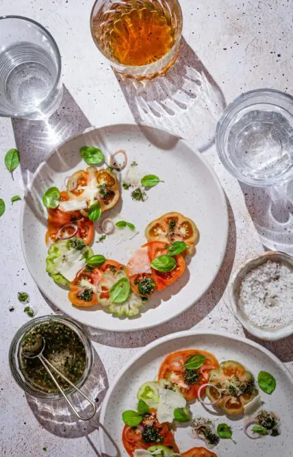 Overhead image of finished heirloom tomato and Red Ruffle basil salads on two white plates, surrounded by glasses of water and a dish of flaky salt