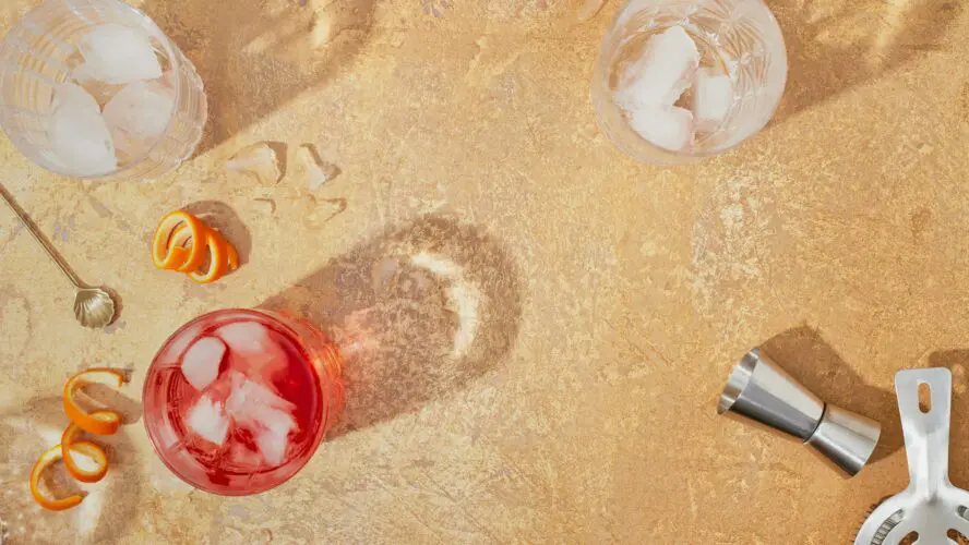 Overhead image of summer drinkscocktails arranged on an outdoor concrete surface surrounded by a shaker, glasses of ice, and a jigger