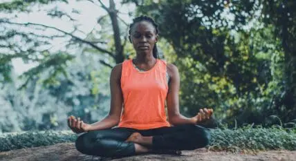 Image of a woman wearing athletic clothing and sitting cross-legged with her eyes closed and her hands resting on her knees, meditating.