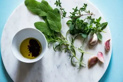 The benefits of flaxseed oil include improved heart health and glowing skin. Image of a round marble slab on a blue background, with a cup of flaxseed oil and balsamic vinegar next to garlic and herbs.