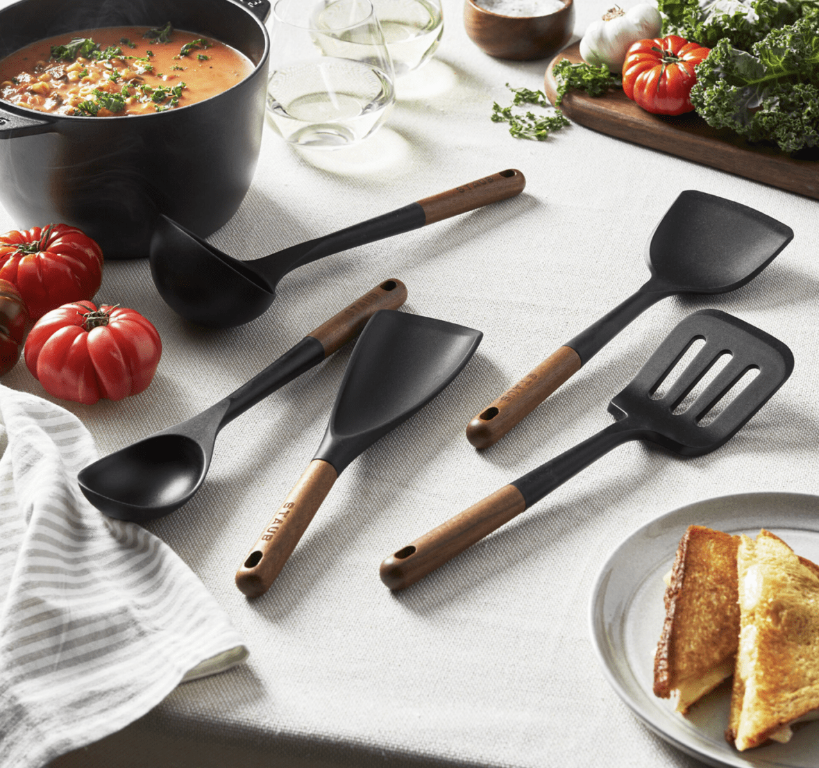 Image of a five-piece set of Staub silicone utensils with acacia wood handles laid out on a white tabletop with a couple of red heirloom tomatoes.
