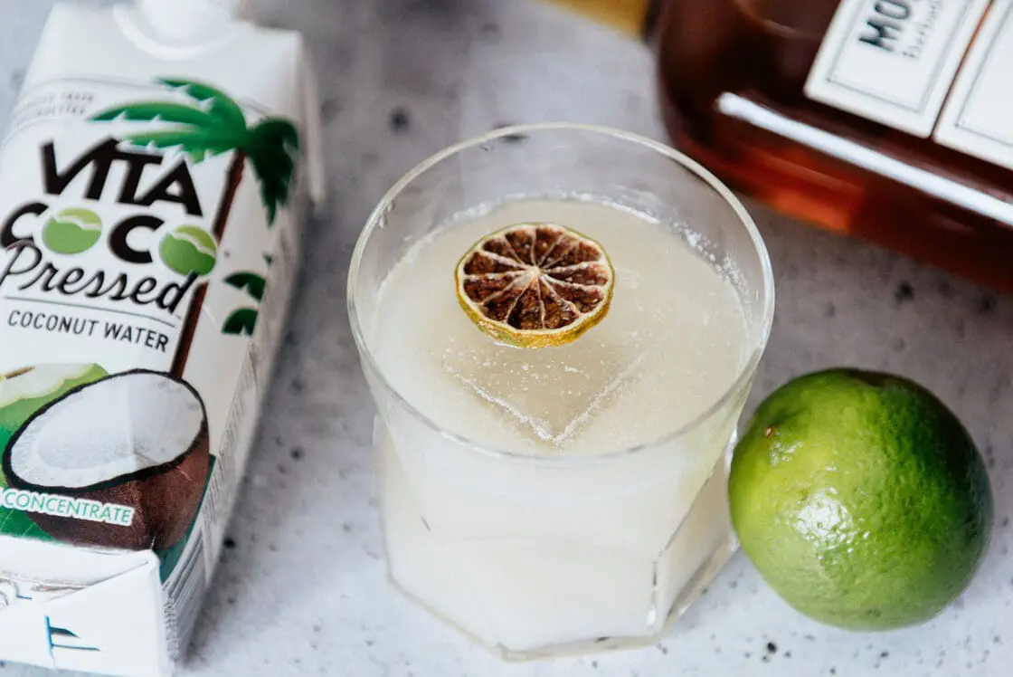 Image of a coconut water and rum cocktail, garnished with some dried blood orange and set next to a lime, a bottle of rum, and a carton of Vita Coco Pressed coconut water.