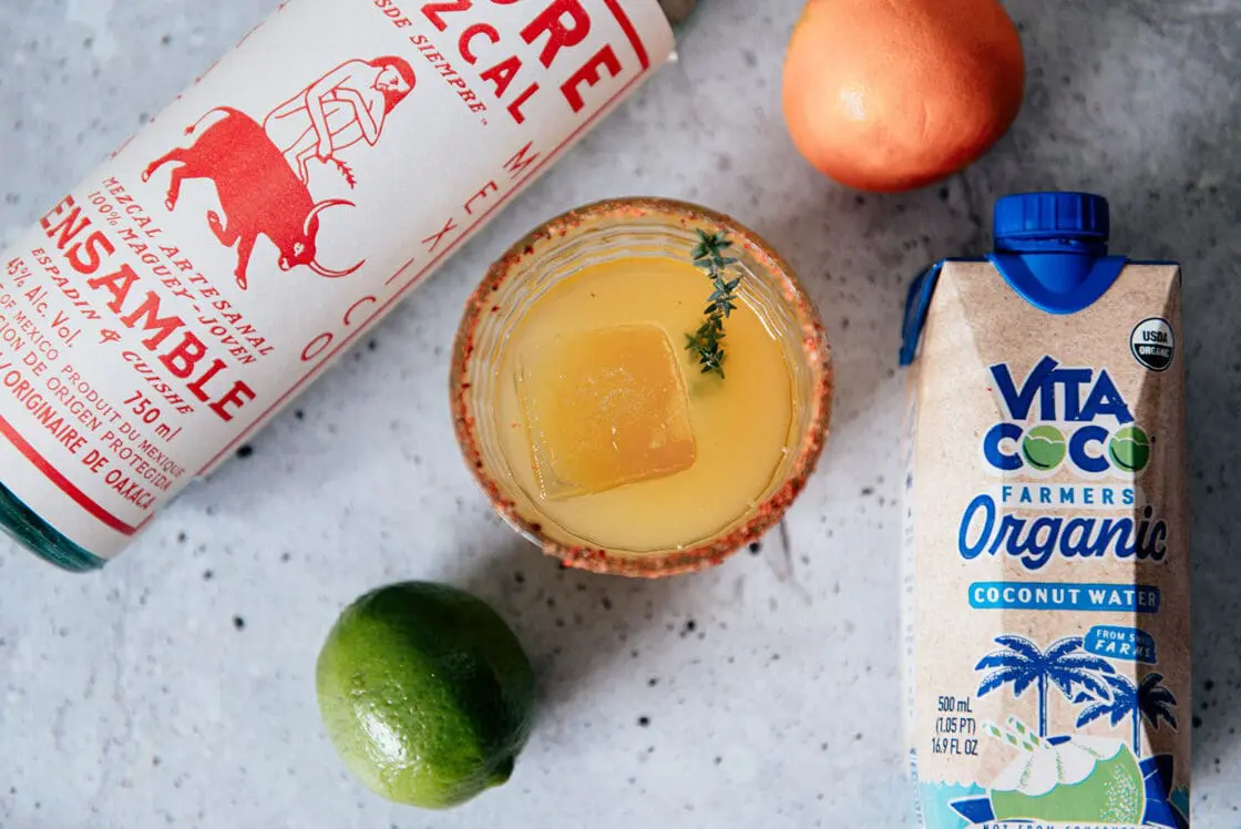 Image of a coconut water mezcalita cocktail with a Tajin rim, set on a marble countertop next to a lime, an orange, a bottle of mezcal, and a carton of Vita Coco Farmer's Organic coconut water.