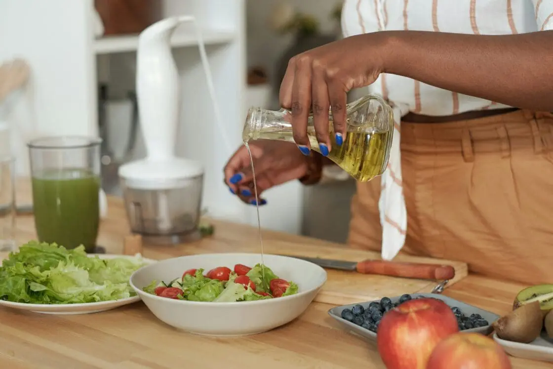 Image of a woman pouring a seed oil from a glass bottle over a fresh green salad on a wooden countertop.