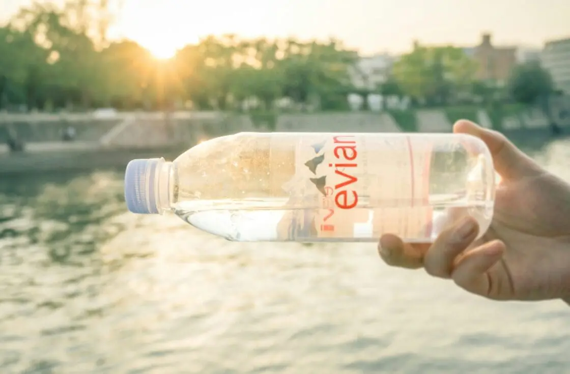 Image of a hand holding an Evian bottle of water on its side, with a small body of water and warm light in the background.