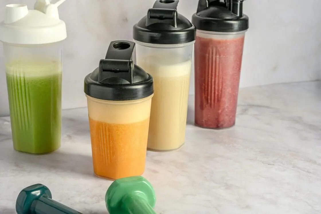 Image of four protein shakes in reusable bottles in various colors, all sitting on a marble tabletop next to a couple of dumbbells.