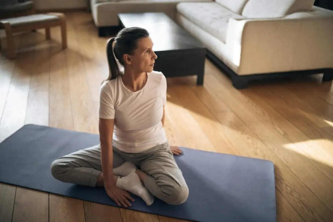 Image of an older woman in a cross-legged position sitting on a yoga mat and looking over her shoulder.