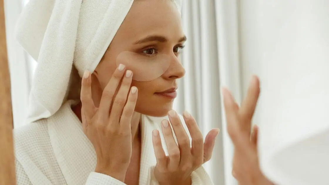 Image of a young woman in a bathrobe and a towel wrapped around her head, looking at herself in the mirror as she applies skincare.