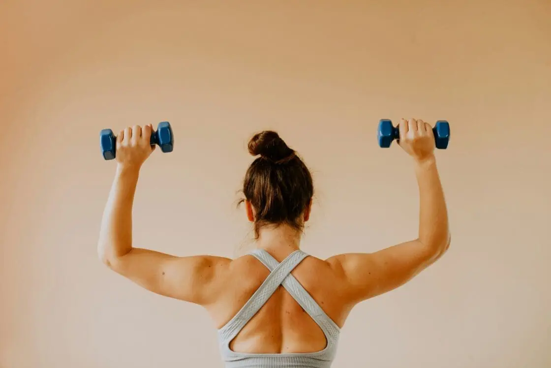 Image of a woman from behind, holding small dumbbells in both hands above her head.