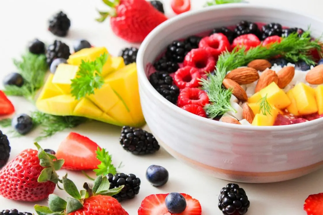 Image of a smoothie bowl featuring berries, mango, almonds, and more, with fruits strewn on the table around the bowl.