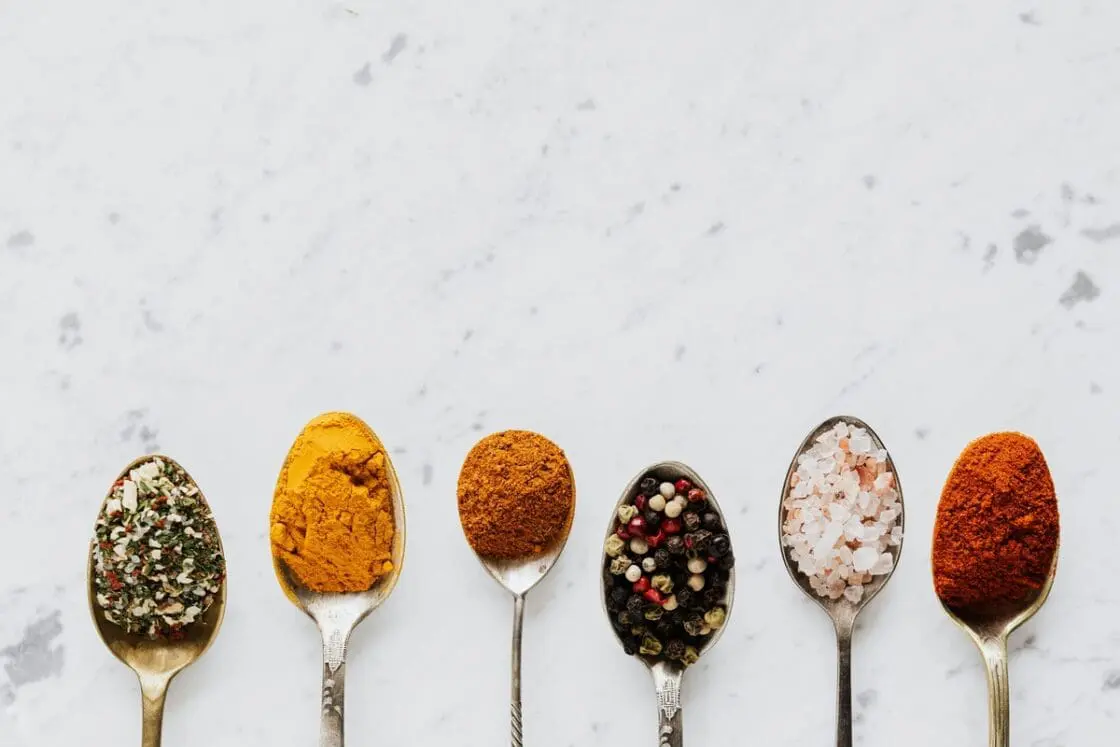 Image of six different spoons on a marble countertop, each filled with a different seasoning, from turmeric and salt to black pepper.
