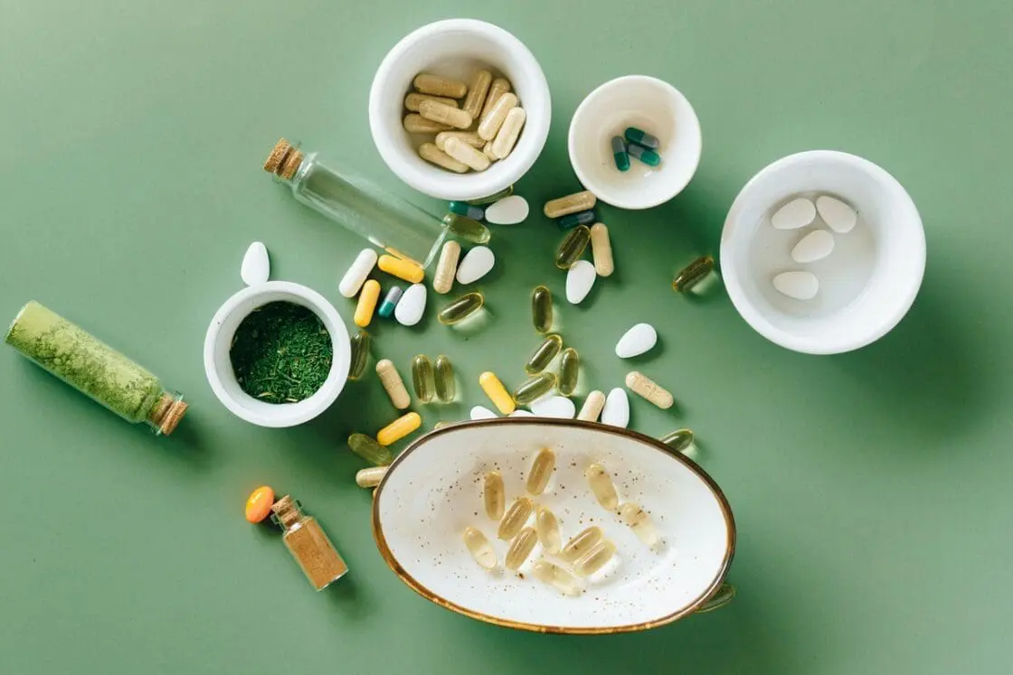 Image of a variety of pills, powders, and supplemental nutrients in various ramekins and cups on a green background.