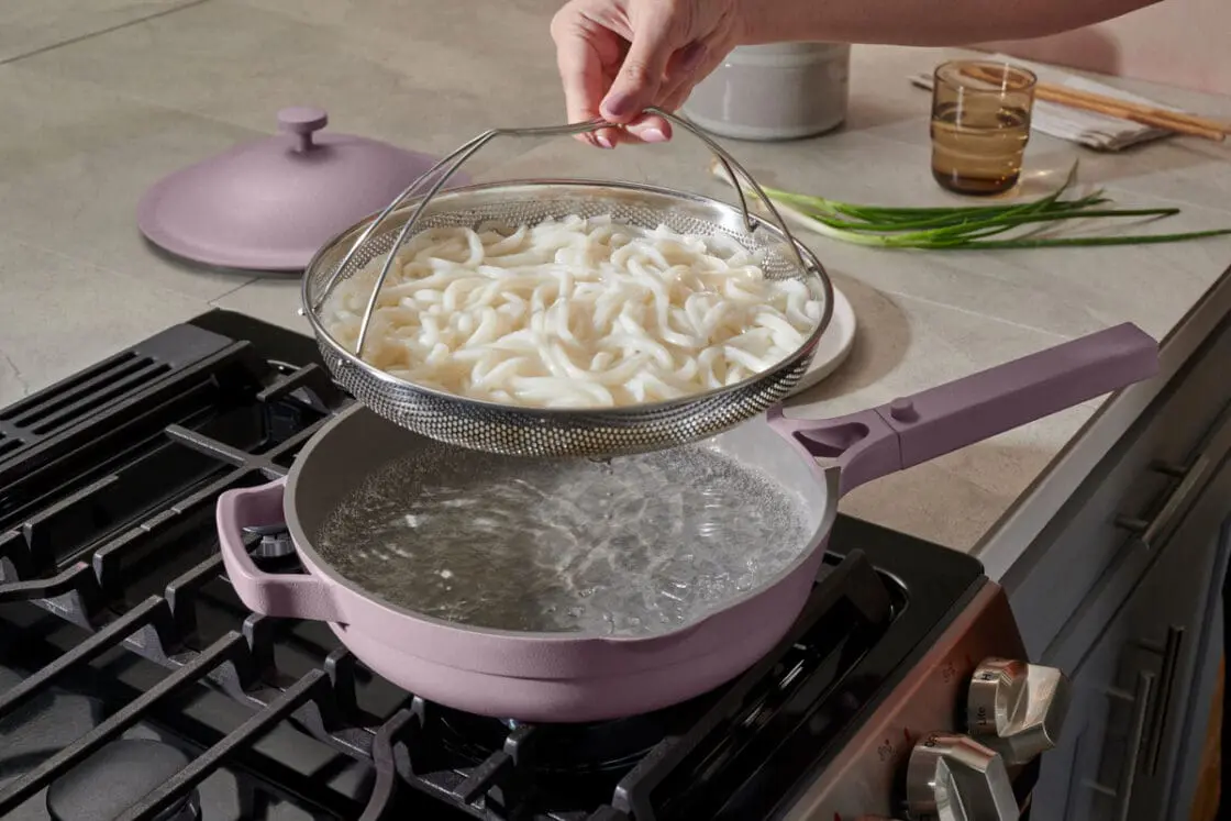 Image of the Our Place Always Pan 2.0 in Lavender, with the included steamer basket full of udon noodles hovering above it on the stovetop.