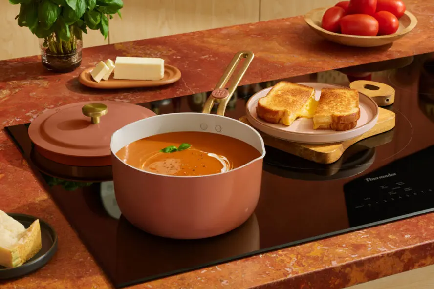 Image of an Our Place Perfect Pan in a gorgeous Terracotta color, shown on a stovetop being used to make tomato soup next to a plate of grilled cheese.