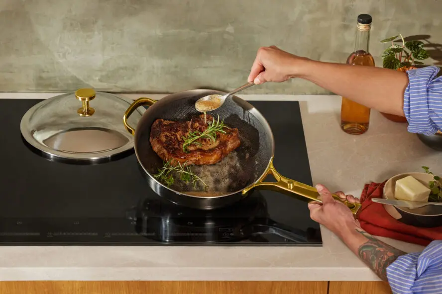 Image of the Titanium Always Pan Pro shown on a stovetop being used to baste a steak, the lid shown on the side.