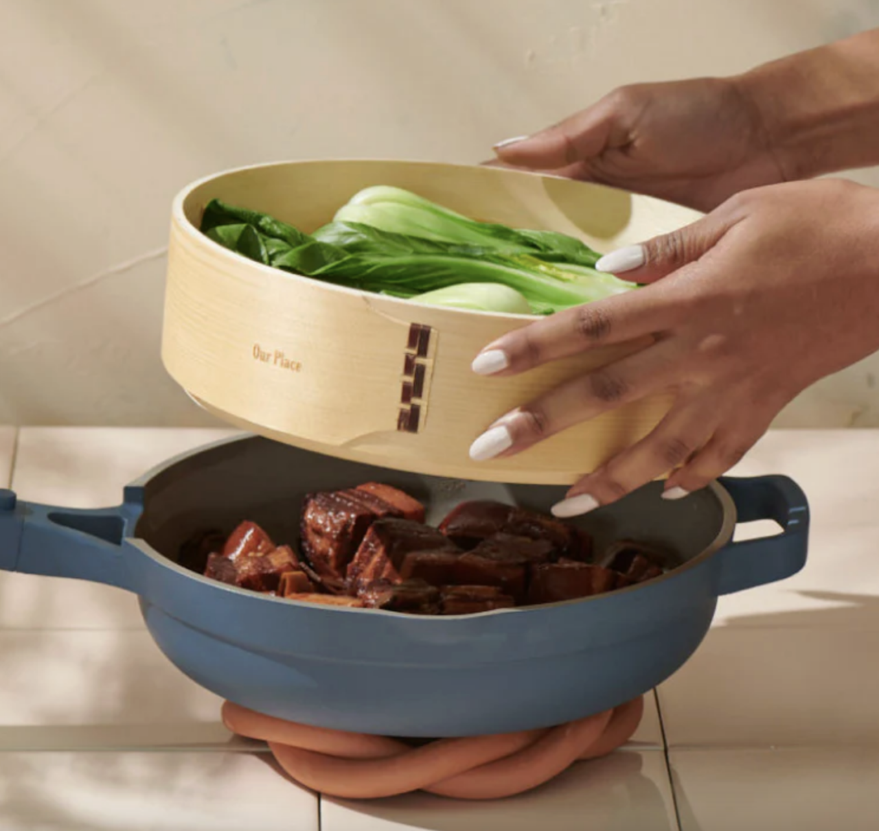 Image of a pair of hands gently placing an Our Place Spruce Steamer Basket full of bok choy over a blue Always Pan.