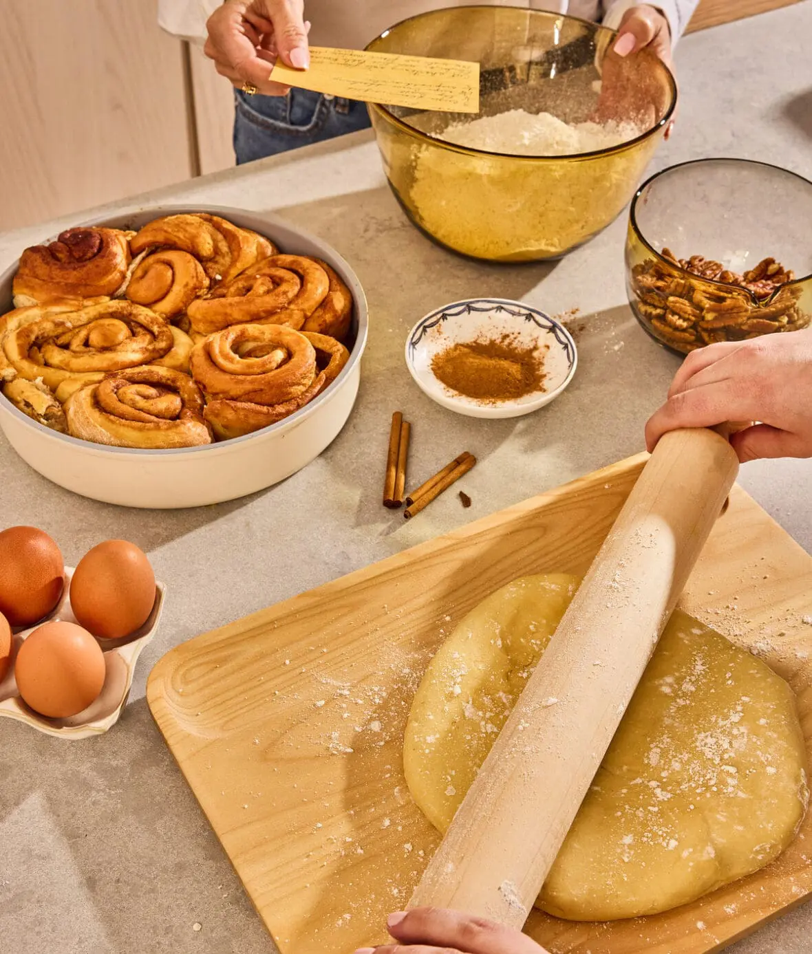 Image of Caraway bakeware and cutting boards in action, with a mother and daughter pair following a recipe card as they roll out dough for cinnamon buns.