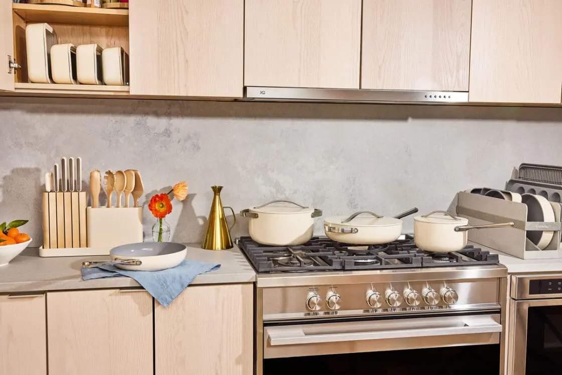 Image of several pieces in the Caraway Cookware set in cream, displayed on a stovetop in a warm kitchen with wooden accents.