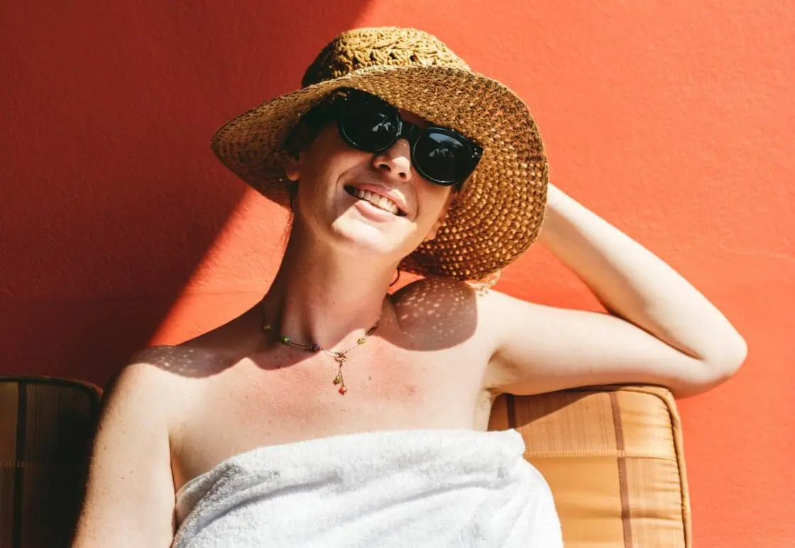 Image of a young woman sitting in the sunshine, wearing a sunhat, towel, and a pair of black sunglasses.