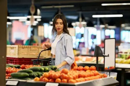 Image of a young woman browsing the fresh produce section at a grocery store, holding a mango in one hand.