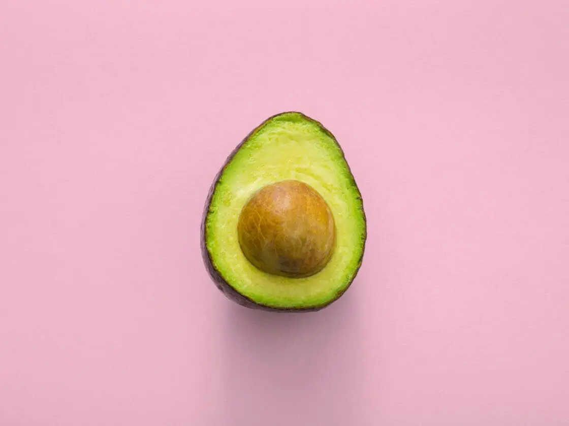 Image of half of a healthy avocado with its pit on a light pink background. Are avocados healthy?