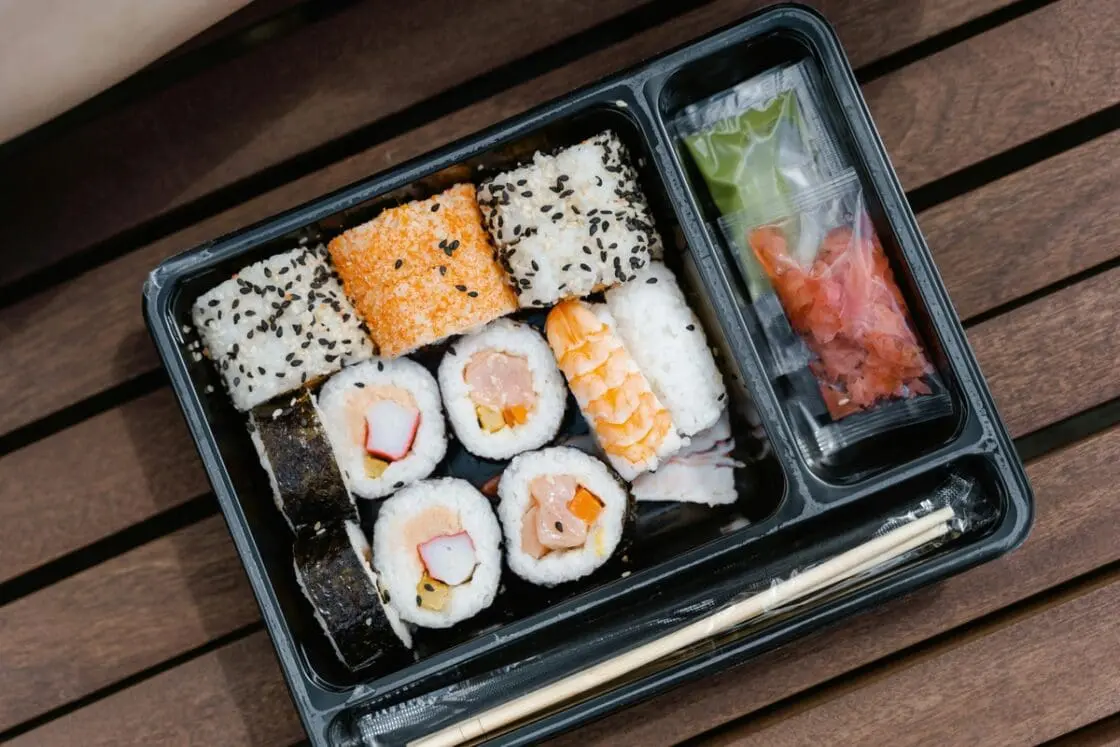 Image of a black plastic takeout container filled with sushi and various condiments, as well as a pair of chopsticks.