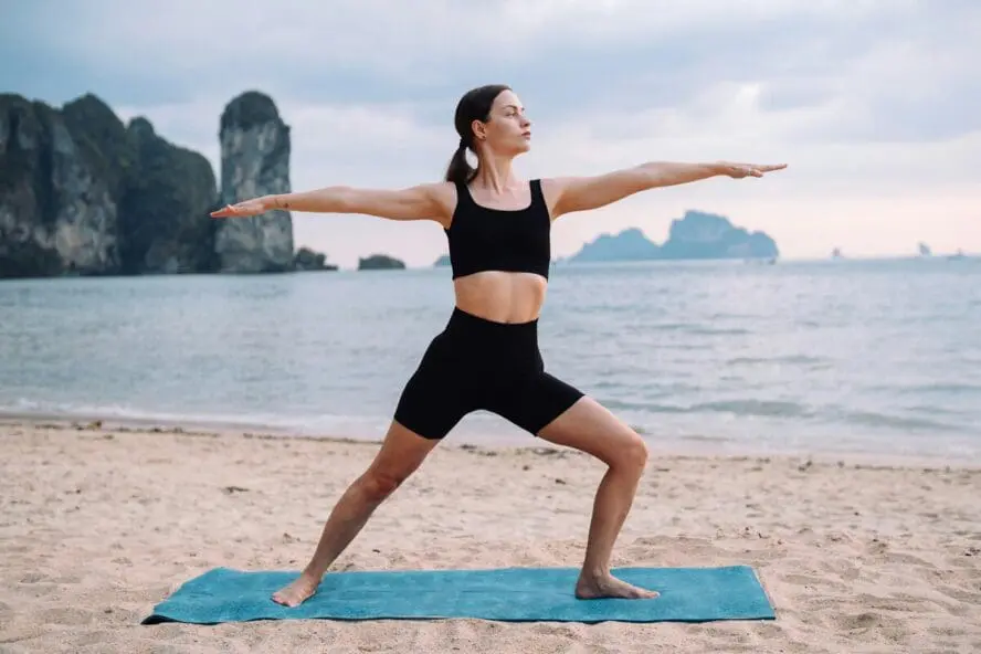 Image of a young woman in a yoga pose on the beach, with water and cliffs behind her.