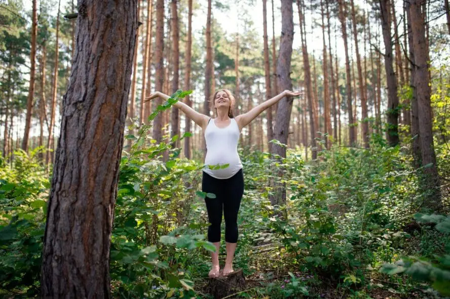 Image of a pregnant woman standing in the forest surrounded by trees, with her arms lifted up above her head.