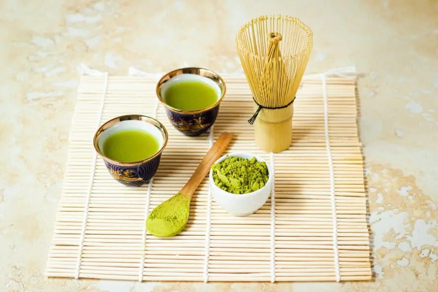 Image of a bamboo mat with two cups of matcha green tea, a bowl of matcha powder, a spoon, and a whisk.