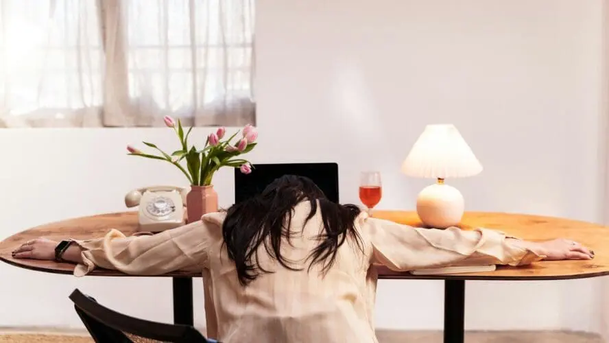 Image of a woman with her arms splayed on a desk in front of her laptop, with her head resting on the table.