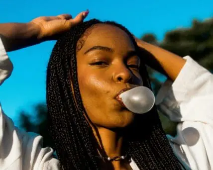 Image of a young black woman with her hands over her head, blowing a bubble with her chewing gum.