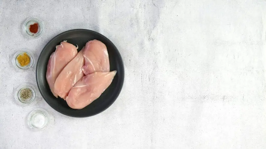 Image of several chicken breasts on a plate alongside several dishes of spices and herbs.