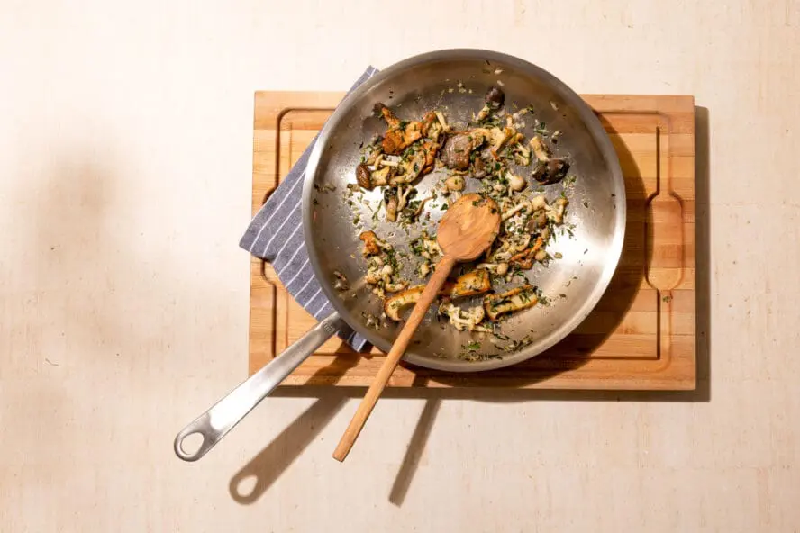 Image of a Made In stainless steel frying pan on a wooden cutting board with cooked mushrooms and a wooden spoon.