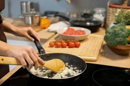 Image of someone cooking onions with a traditional nonstick pan on a glass stovetop, with tomatoes on a cutting board next to them.